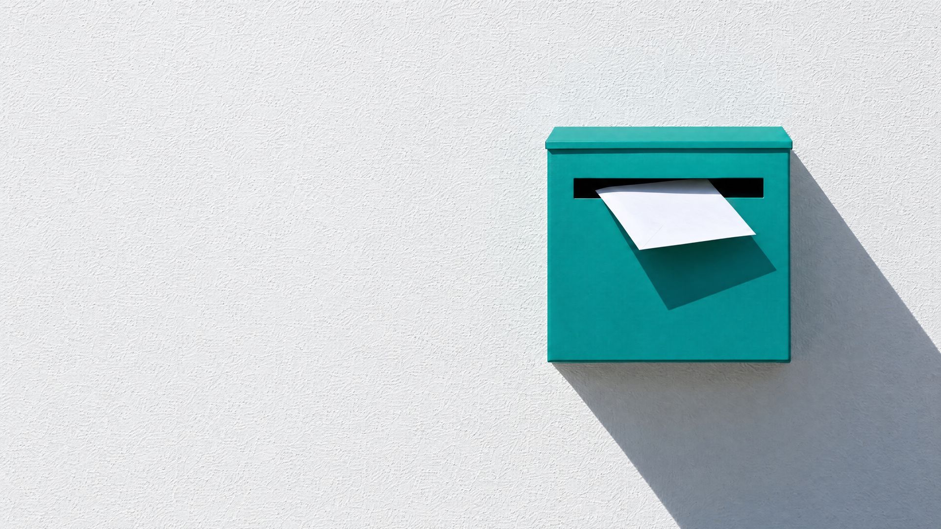 A flat teal letterbox on a sunlit white wall, with a white envelope halfway through its slot.
